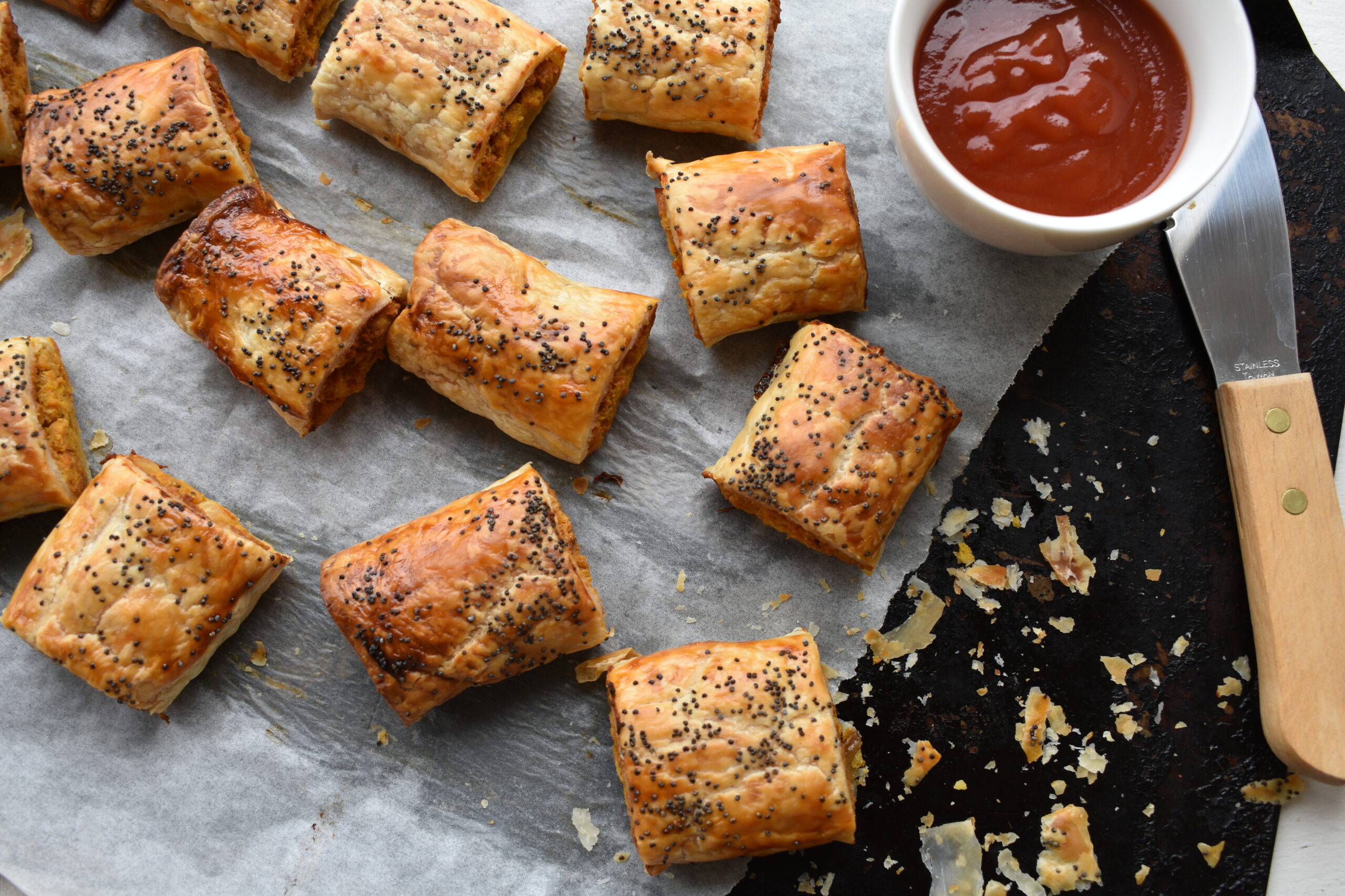 Vegan Sausage Rolls with Poppy seeds. On baking tray with tomato sauce and knife